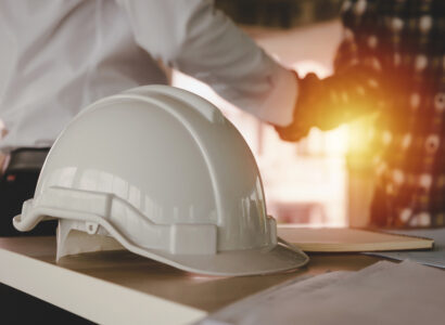 A white hard hat sits on a table in the foreground while two people shake hands in the blurred background.