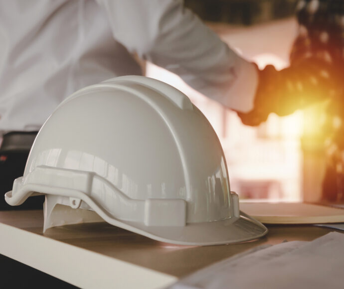 A white hard hat sits on a table in the foreground while two people shake hands in the blurred background.