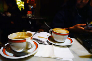 Two coffee drinks in mugs atop saucers on a table with an open notebook and laptop.