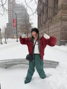 Employee Charlotte wearing a red jacket and green pants standing in snow up to her knees, giving two thumbs up and a big smile.
