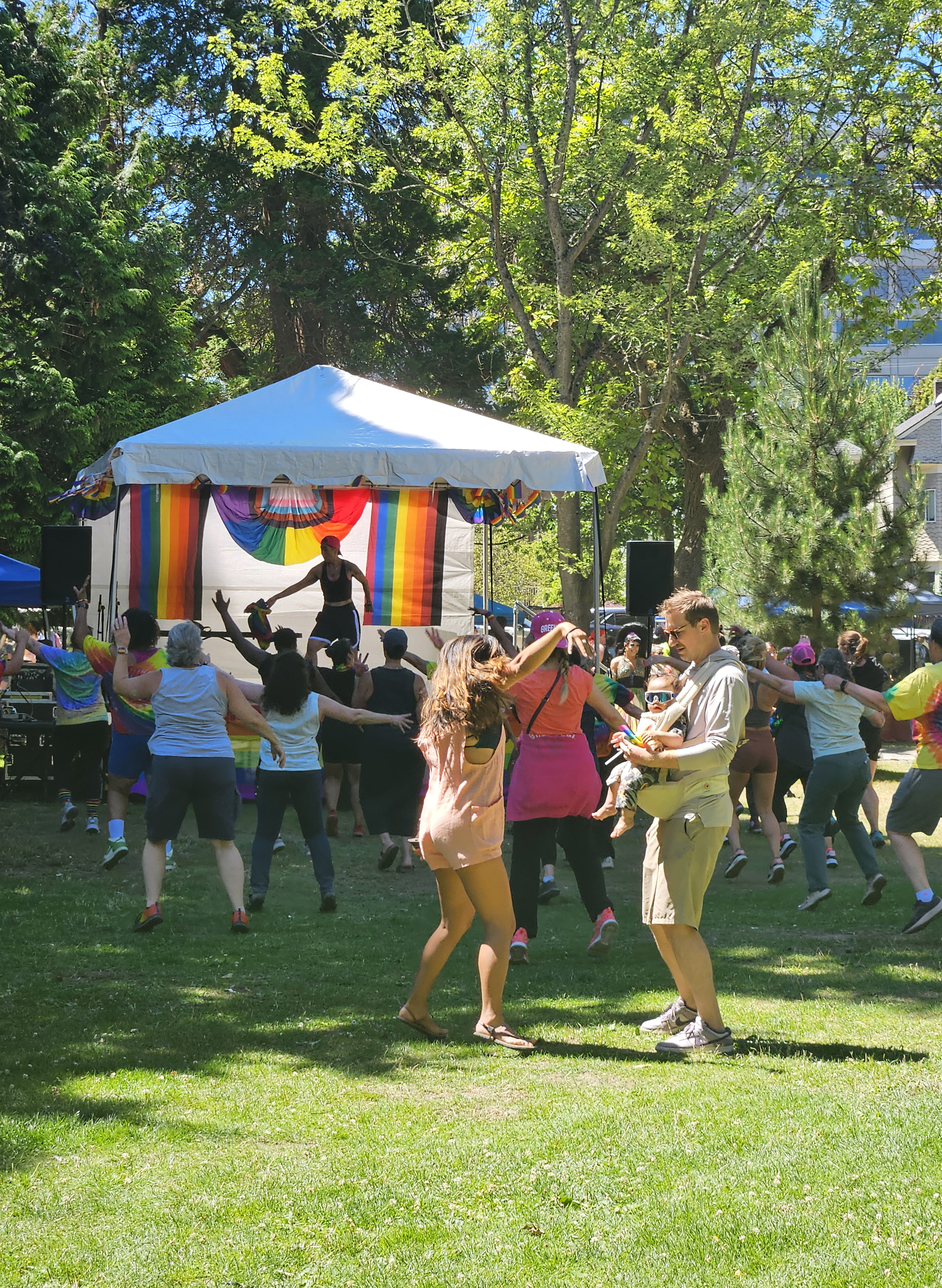A mother dances with her partner that is holding their baby, donning sunglasses and a small Pride flag as a large group of people dance behind them.