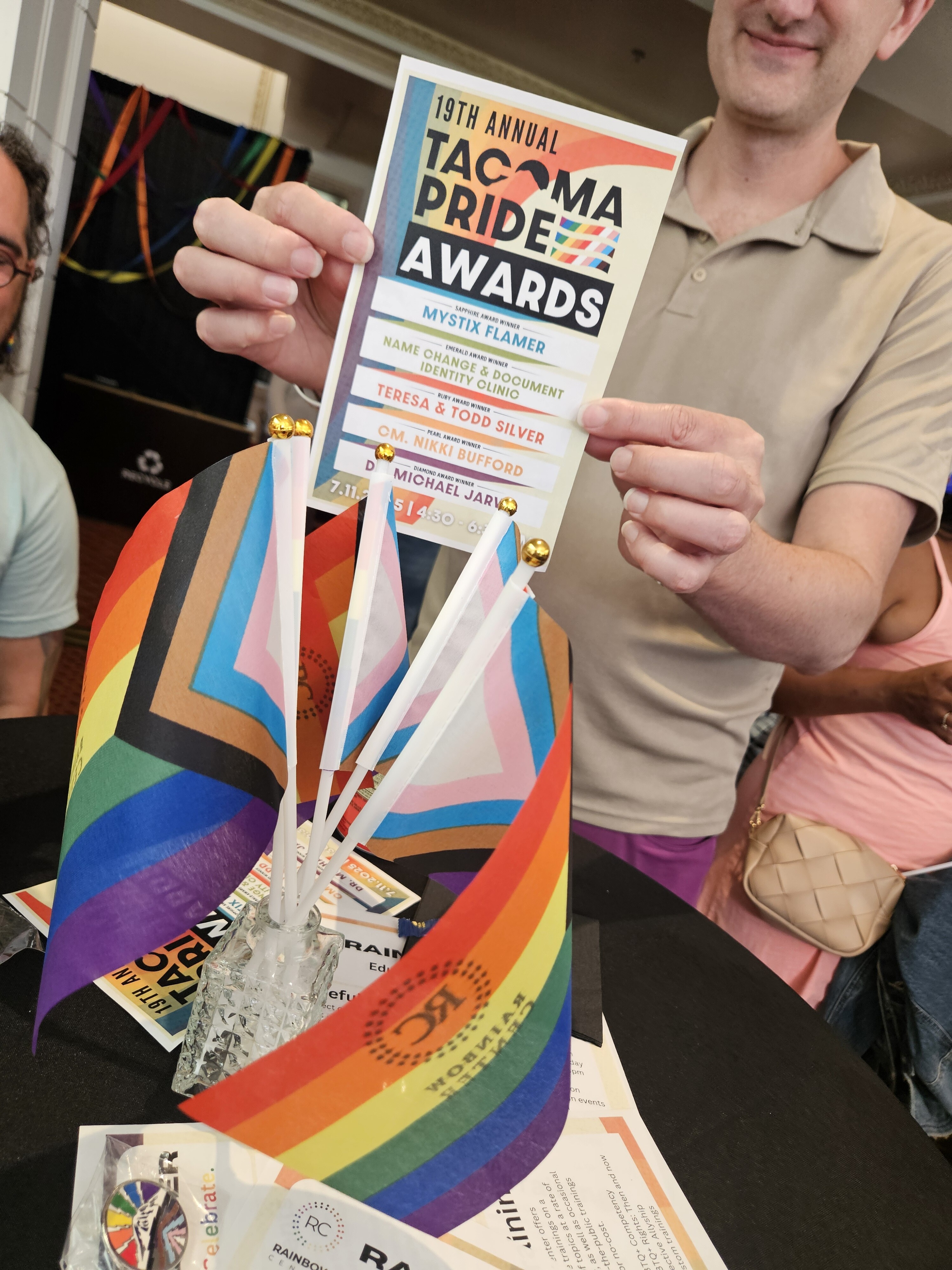 A program for Annual Tacoma Pride Awards is held up above a table with Pride flags and pins