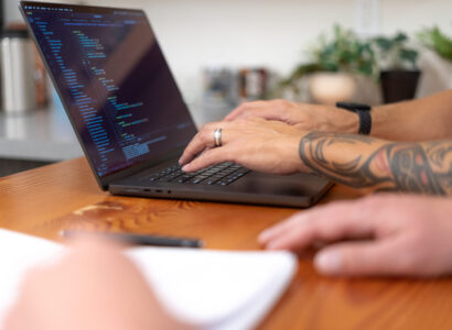 Two SiteCrafting employees collaborating. One employee is typing on a laptop. The other employee is seated beside them, writing notes in a spiral notebook.