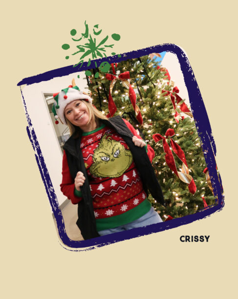 SiteCrafting employee Crissy wearing a red Grinch sweater, a black vest and a festive elf hat, smiling next to a Christmas tree.
