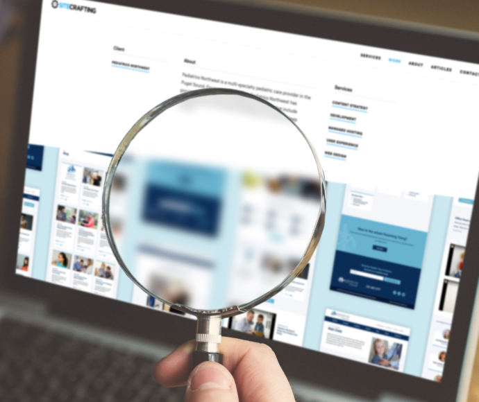 A bearded man holding a magnifying glass in front of a laptop screen.