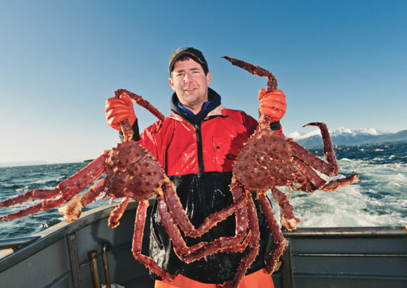 Man on boat holding very large crab in each hand on a sunny day.