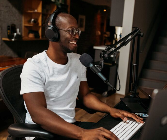 African American man sitting at a computer wearing headphones and talking into a microphone.