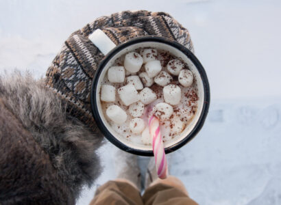 Person stand in snow holding a mug with mittens on