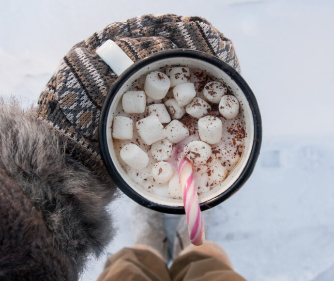 Person stand in snow holding a mug with mittens on