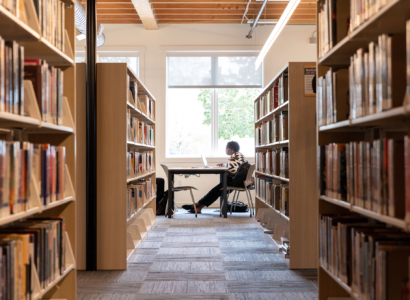 A person sitting at a desk working on a laptop, framed between tall rows of library bookshelves filled with books.