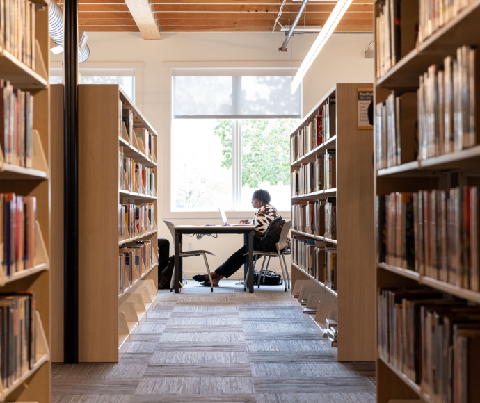 A person sitting at a desk working on a laptop, framed between tall rows of library bookshelves filled with books.
