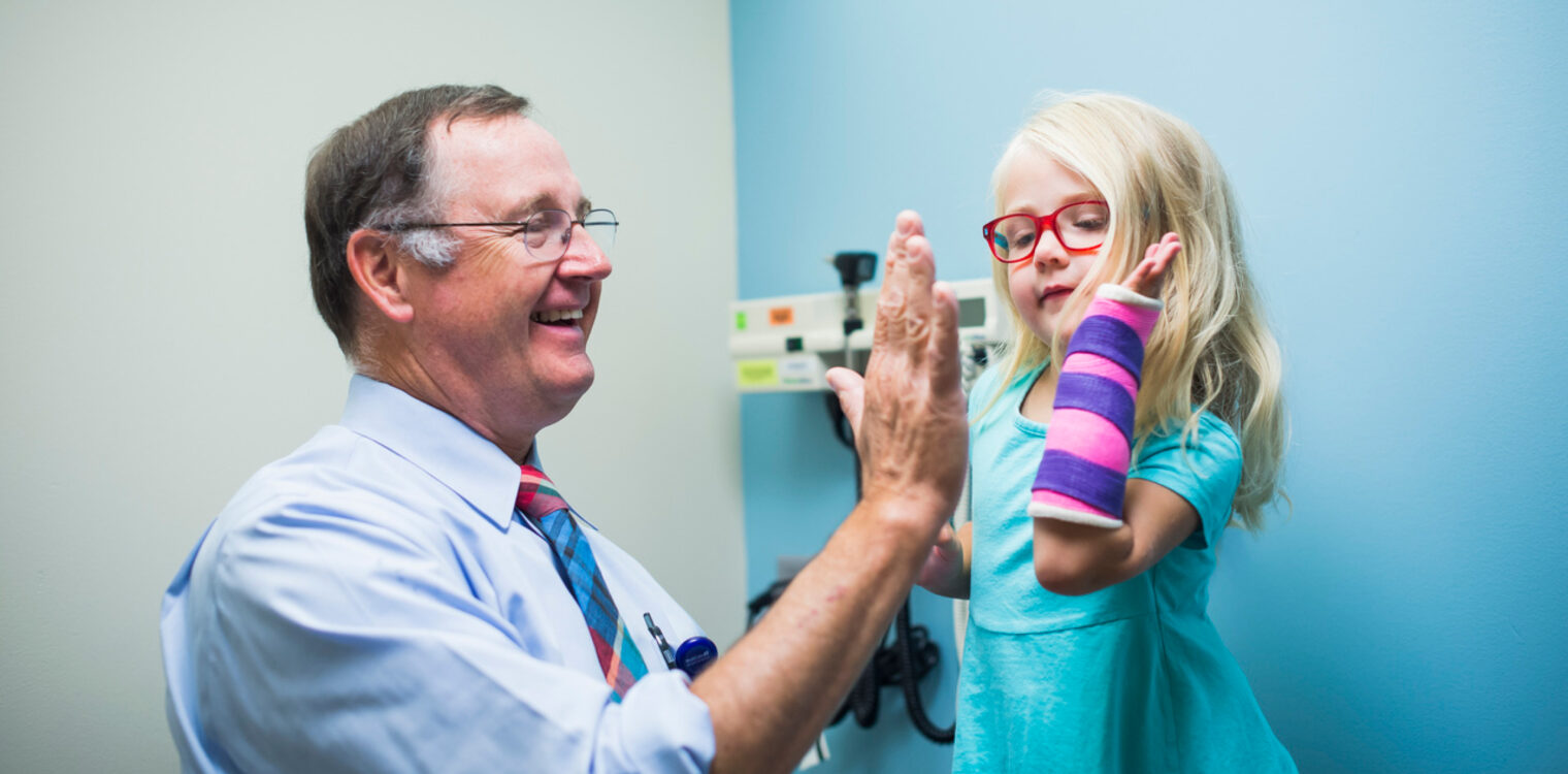 Medical professional in a light blue shirt and red-striped tie interacting with a child wearing red glasses and a colorful arm cast. The child reaches out with the casted arm, possibly for a high-five or exam, in a medical setting with equipment in the background.