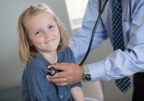 A small blond girl smiling while a doctor listens to her heartbeat with a stethoscope.