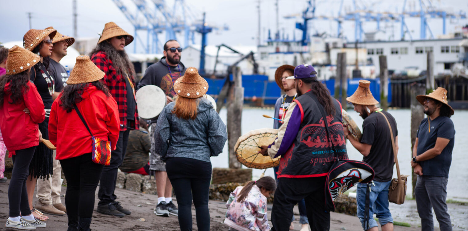 Members of the Puyallup Tribe of Indians standing in a circle on the beach holding drums