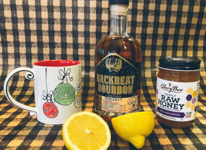 Mug, bourbon whiskey, raw honey and a fresh cut lemon sitting on a counter.