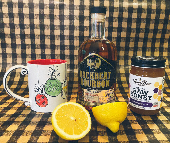 Mug, bourbon whiskey, raw honey and a fresh cut lemon sitting on a counter.