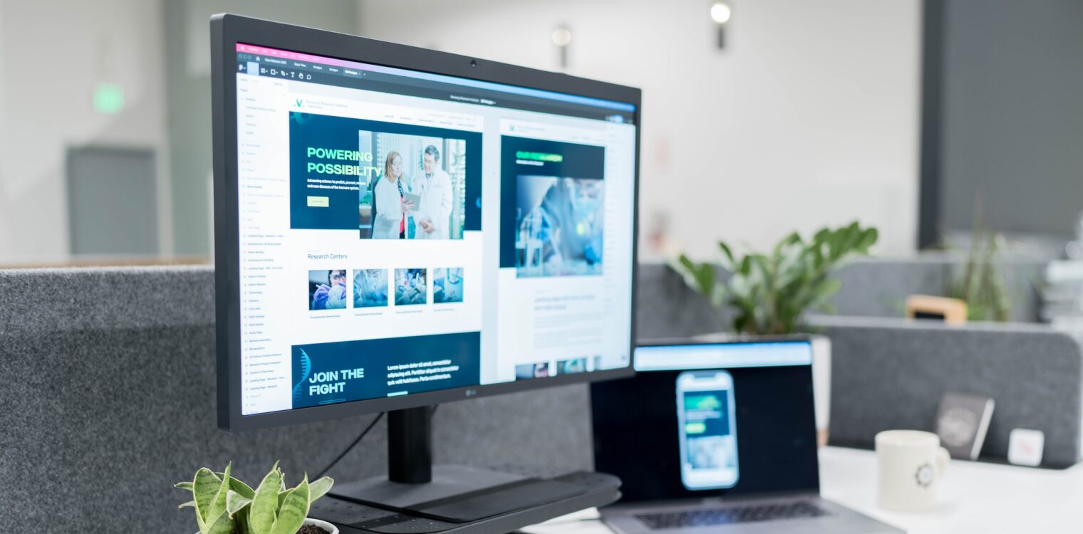 A monitor and laptop on a desk in an office. The monitor displays a website with text that reads 